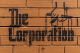 Stencil graffiti art on a brick wall features the words 'The Corporation' with an image of hands manipulating puppet strings.