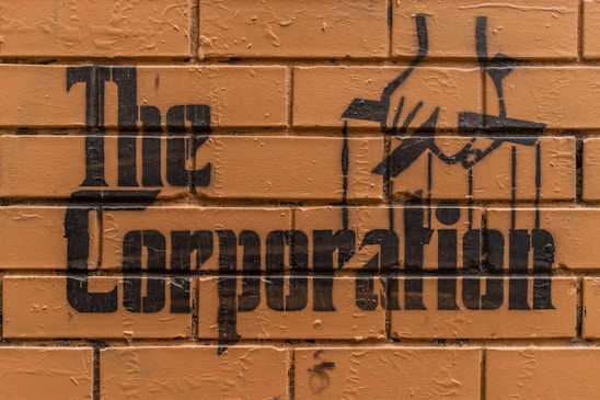 Stencil graffiti art on a brick wall features the words 'The Corporation' with an image of hands manipulating puppet strings.