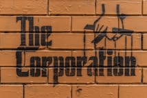 Stencil graffiti art on a brick wall features the words 'The Corporation' with an image of hands manipulating puppet strings.