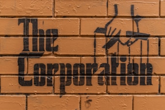 Stencil graffiti art on a brick wall features the words 'The Corporation' with an image of hands manipulating puppet strings.