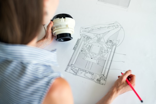A person is closely examining a technical drawing on a large sheet of paper with detailed diagrams. They are holding a red pencil and a white mechanical object, suggesting they may be comparing the object to the drawing. The person is wearing a blue and white striped shirt.