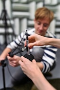 A person with a focused expression is kneeling and appears to be assisting in adjusting or connecting a microphone. Another pair of hands is seen securing a foam windscreen onto the microphone. The setting is likely a recording or acoustic testing room with soundproofing material visible in the background.