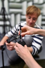 A person with a focused expression is kneeling and appears to be assisting in adjusting or connecting a microphone. Another pair of hands is seen securing a foam windscreen onto the microphone. The setting is likely a recording or acoustic testing room with soundproofing material visible in the background.