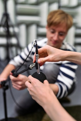 A person with a focused expression is kneeling and appears to be assisting in adjusting or connecting a microphone. Another pair of hands is seen securing a foam windscreen onto the microphone. The setting is likely a recording or acoustic testing room with soundproofing material visible in the background.