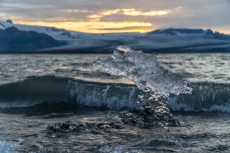 A clear, sculptural piece of ice shaped by the water stands prominently in front of a gentle wave. In the background, a dramatic landscape of distant mountains is partially illuminated by a setting or rising sun, casting a warm glow over the scene.