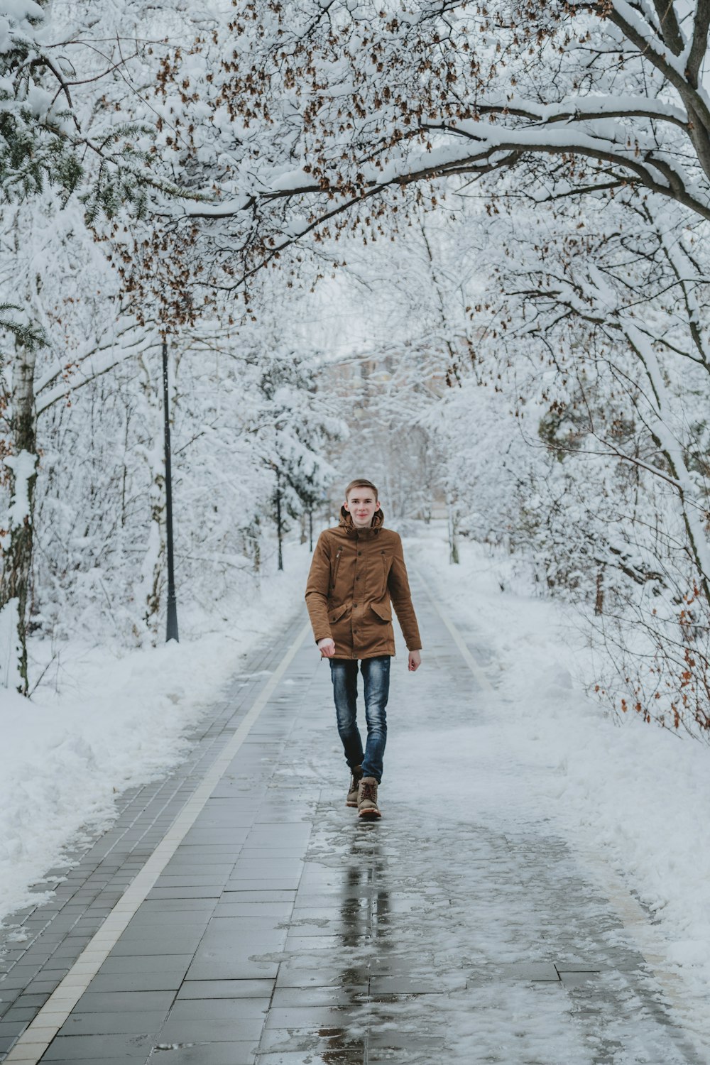 woman in brown coat walking on snow covered pathway between trees during daytime