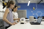Two women are engaged in a laboratory setting. They are working on an electronic project involving a circuit board and a small device placed on the table. The workspace is equipped with modern technology, and there is a laptop in use, likely for coding or data analysis. The background features blue walls with scientific patterns and equipment.