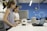 Two women are engaged in a laboratory setting. They are working on an electronic project involving a circuit board and a small device placed on the table. The workspace is equipped with modern technology, and there is a laptop in use, likely for coding or data analysis. The background features blue walls with scientific patterns and equipment.