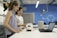 Two women are engaged in a laboratory setting. They are working on an electronic project involving a circuit board and a small device placed on the table. The workspace is equipped with modern technology, and there is a laptop in use, likely for coding or data analysis. The background features blue walls with scientific patterns and equipment.