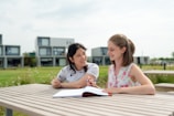 Two people are sitting at a wooden picnic table in an outdoor area, engaged in a conversation or study session. Both seem relaxed and are smiling, with one of them holding a pen and an open notebook in front of them. In the background, there are modern, cube-like buildings and a well-maintained grassy area.