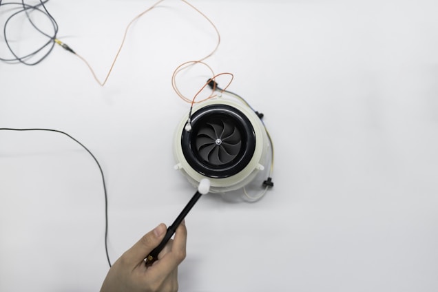 A hand is holding a small device with an attached probe near an electrical fan or turbine surrounded by wires on a clean white surface. The fan is circular with multiple blades, and several wires are connected to it.