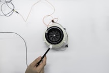 A hand is holding a small device with an attached probe near an electrical fan or turbine surrounded by wires on a clean white surface. The fan is circular with multiple blades, and several wires are connected to it.