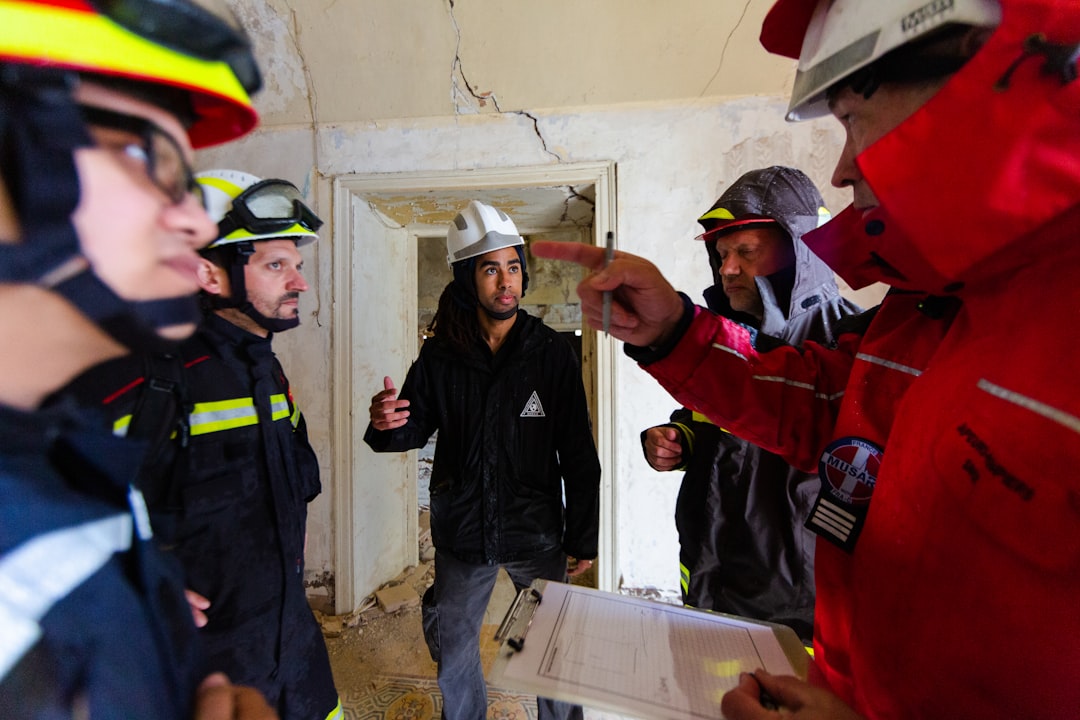 3 men in red and black jacket standing beside white wall during daytime, Structural engineers in disaster relief training