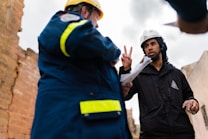 Two individuals wearing safety helmets engage in a discussion at a construction site. One person appears to be gesturing with two fingers, holding a piece of paper, while the other is in a blue jacket with reflective stripes.