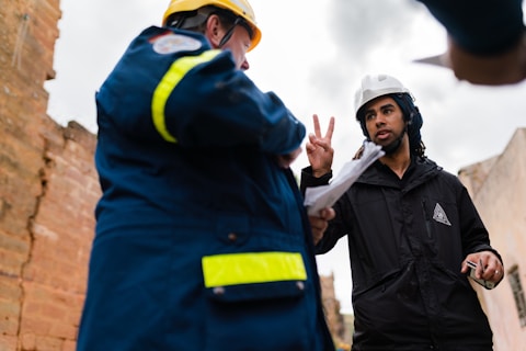 Two individuals wearing safety helmets engage in a discussion at a construction site. One person appears to be gesturing with two fingers, holding a piece of paper, while the other is in a blue jacket with reflective stripes.