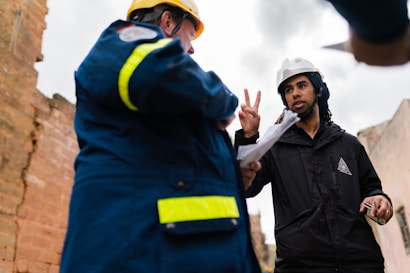 Two individuals wearing safety helmets engage in a discussion at a construction site. One person appears to be gesturing with two fingers, holding a piece of paper, while the other is in a blue jacket with reflective stripes.