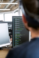 A person is focused on a computer screen displaying a large amount of code. There are two monitors on the desk, one of which has code while the other appears less clear. The scene is set in an office environment with a ceiling featuring exposed pipes and large windows in the background allowing natural light to shine through.
