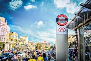 A busy urban street bustling with cars, motorcycles, and people. The scene includes buildings with various signs and advertisements on the left, and a tall sign marked 'Hazrat Ganj' on the right. The sky is clear with a few clouds.