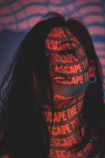 A person with long, dark hair stands in a shadowy environment. Red letters spelling 'ESCAPE THE' are projected across their face and neck in a repeating pattern, creating a mysterious and artistic effect. The background features a distorted pattern of red and blue wavy lines.