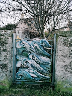 A decorative metal gate featuring an artistic wave pattern stands between two stone pillars. Behind the gate, a barren tree is visible, with branches reaching outwards in various directions. In the background, a traditional house is partially obscured by the tree, giving an impression of an older, possibly rural setting.
