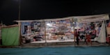 A makeshift market stall brightly lit at night displays a wide variety of colorful folded textiles stacked in organized piles. Two people stand in front of the stall, one of them appearing to interact with the vendor, while chairs are placed casually next to them.
