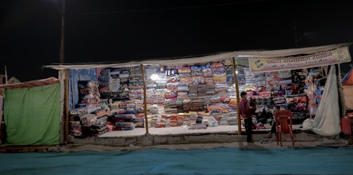 A makeshift market stall brightly lit at night displays a wide variety of colorful folded textiles stacked in organized piles. Two people stand in front of the stall, one of them appearing to interact with the vendor, while chairs are placed casually next to them.