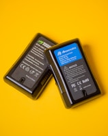 Two black lithium-ion battery packs are placed against a vibrant yellow background. The battery on the right has a blue label with technical specifications and safety notices, while the battery on the left features a black and green label with similar information.
