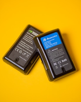 Two black lithium-ion battery packs are placed against a vibrant yellow background. The battery on the right has a blue label with technical specifications and safety notices, while the battery on the left features a black and green label with similar information.