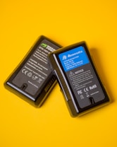 Two black lithium-ion battery packs are placed against a vibrant yellow background. The battery on the right has a blue label with technical specifications and safety notices, while the battery on the left features a black and green label with similar information.