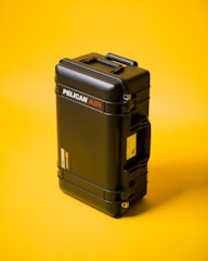 A black hard-shell protective case labeled 'Pelican Air' stands upright against a bright yellow background. The case features robust construction with secure clasps and a handle on the side.