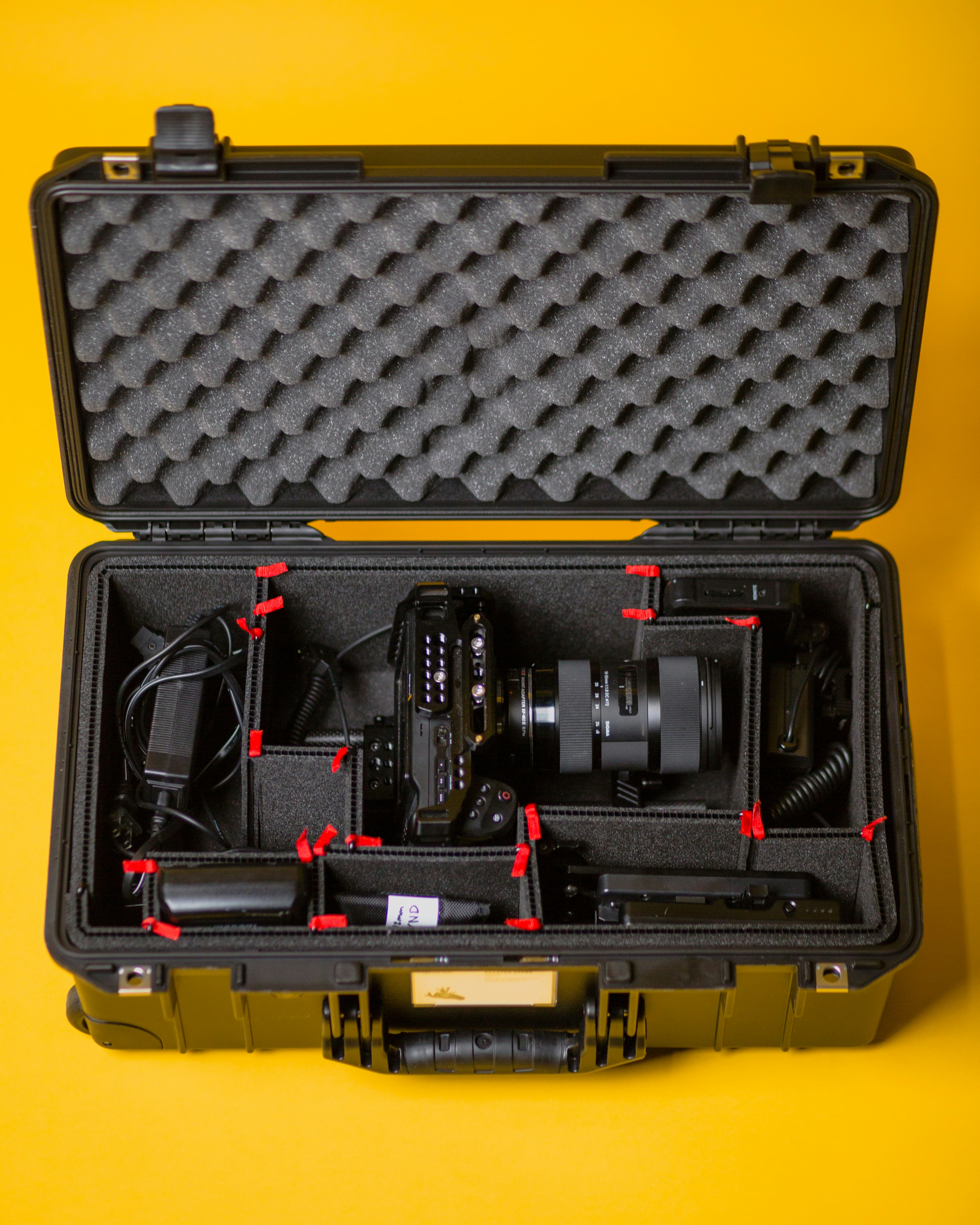 Open black hard case on a yellow backdrop, its foam-lined lid up and a camera, lens, cables and batteries sitting in cut-foam compartments marked with small red tags