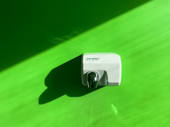 A hand dryer is mounted on a vibrant green wall. The hand dryer is white with a shiny metallic nozzle and casts a distinct shadow on the wall, indicating a light source from the upper left corner.