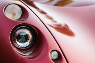A close-up view of a vintage or classic car's headlight assembly. The image focuses on the curved, glossy red bodywork and the round headlight with a reflective surface. The smoothness and shine of the car's paint suggest it is well-maintained. Lighting creates soft reflections across the surface.