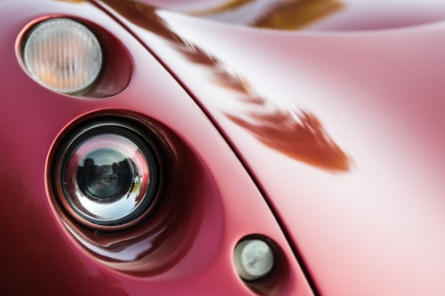 A close-up view of a vintage or classic car's headlight assembly. The image focuses on the curved, glossy red bodywork and the round headlight with a reflective surface. The smoothness and shine of the car's paint suggest it is well-maintained. Lighting creates soft reflections across the surface.