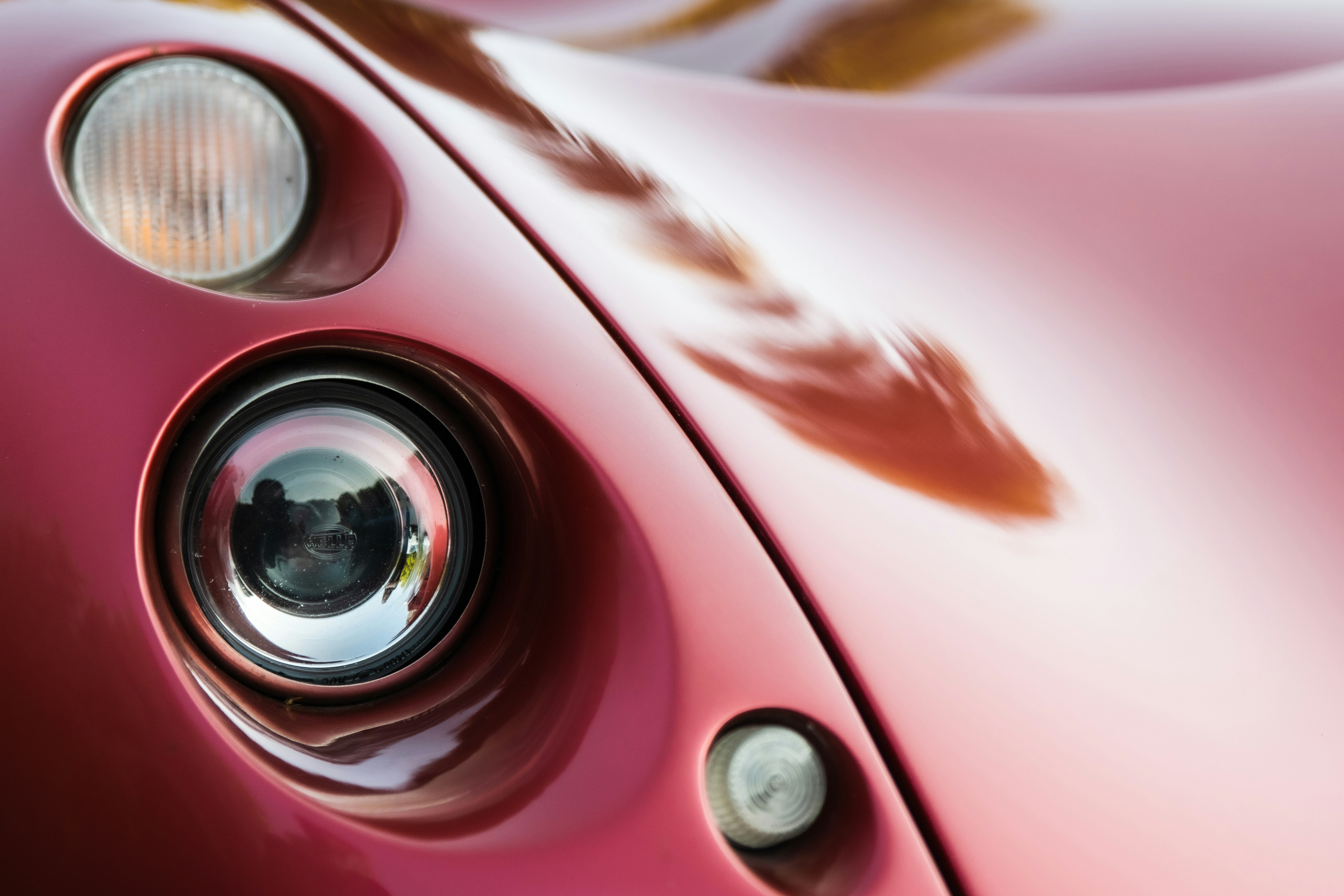 A close-up view of a vintage or classic car's headlight assembly. The image focuses on the curved, glossy red bodywork and the round headlight with a reflective surface. The smoothness and shine of the car's paint suggest it is well-maintained. Lighting creates soft reflections across the surface.