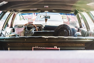 The interior of a vintage car with black leather seats is shown, viewed from the trunk area. On the back seats are items adorned with the American flag pattern, including a backpack and a hat. The rear window shows a parking lot with various vehicles visible in the background.