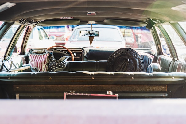 The interior of a vintage car with black leather seats is shown, viewed from the trunk area. On the back seats are items adorned with the American flag pattern, including a backpack and a hat. The rear window shows a parking lot with various vehicles visible in the background.