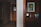 A dimly lit hallway in a hotel or apartment, featuring a wooden door with a sign and a side table with a small coffee maker, a bottle of water, and a few booklets. A piece of modern art depicting green leaves is hung on the wooden wall panel above the counter.