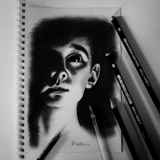 A realistic pencil drawing of a person's face is depicted on a spiral-bound sketchpad. The portrait captures a dramatic use of light and shadow, enhancing the subject's gaze and facial features. Several graphite pencils are placed beside the sketchpad, showcasing different shades and emphasizing the monochromatic theme.