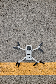 A drone with black propellers is positioned on a textured asphalt surface with a prominent yellow line running horizontally beneath it.