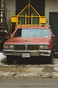 A vintage red car, likely a Pontiac, is parked in front of a garage. The car is fitted with a prominent yellow structure on its roof, possibly for transportation or utility purposes. The license plate indicates Ontario, and dirty snow surrounds the vehicle, suggesting a winter or early spring setting. The background shows a garage door and various tools or equipment stacked nearby.