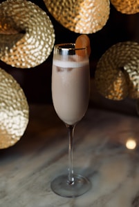 A tall glass filled with a creamy beverage is garnished with a thin slice of citrus. The glass sits on a smooth marbled surface with golden, textured decorations in the background, creating a luxurious and sophisticated atmosphere.
