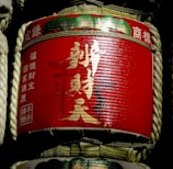 A traditional sake barrel wrapped in straw, featuring bright red labels with gold and white Japanese kanji characters. The barrel is cylindrical, with ropes tied around it for support.