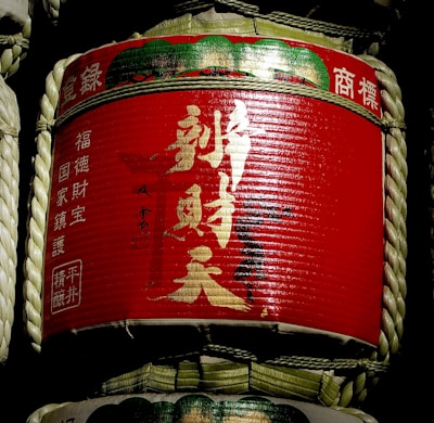 A traditional sake barrel wrapped in straw, featuring bright red labels with gold and white Japanese kanji characters. The barrel is cylindrical, with ropes tied around it for support.