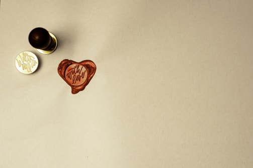 A wax seal with a heart shape and the words 'with love' is stamped on a light-colored surface. A handle, likely part of a seal stamp, rests nearby along with the metal stamp component showing the inverted phrase.