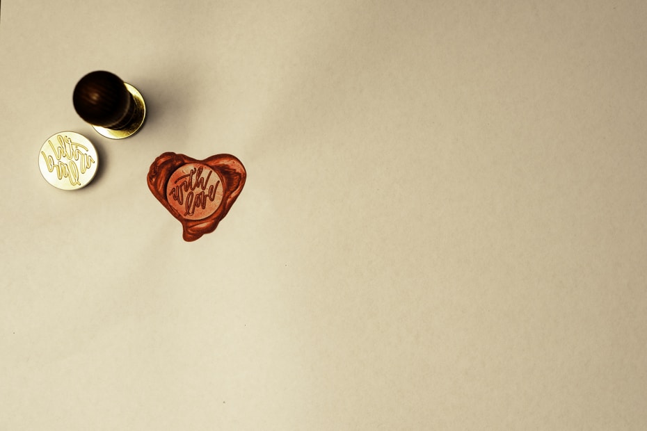 A wax seal with a heart shape and the words 'with love' is stamped on a light-colored surface. A handle, likely part of a seal stamp, rests nearby along with the metal stamp component showing the inverted phrase.