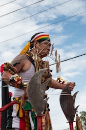 A person dressed in colorful traditional attire participates in a cultural or religious parade. The individual is adorned with ornaments, beads, and wears a multicolored headband. They appear to be engaged in a ritual or performance involving large, curved metal objects and skewers through the cheeks. The sky is partly cloudy and power lines are visible in the background.