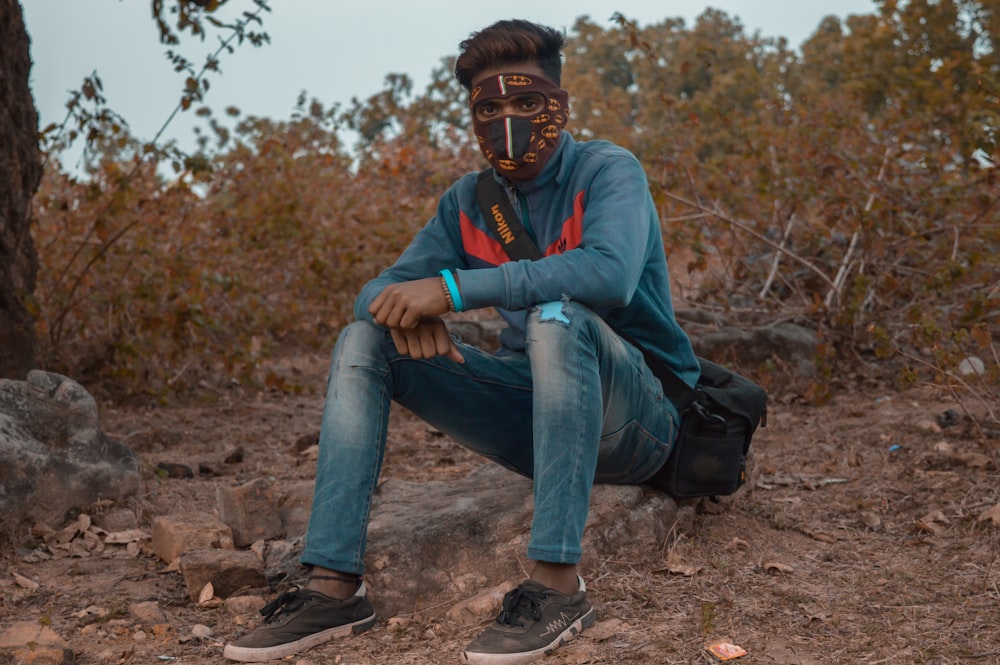 man in blue and red hoodie and blue denim jeans sitting on rock during daytime