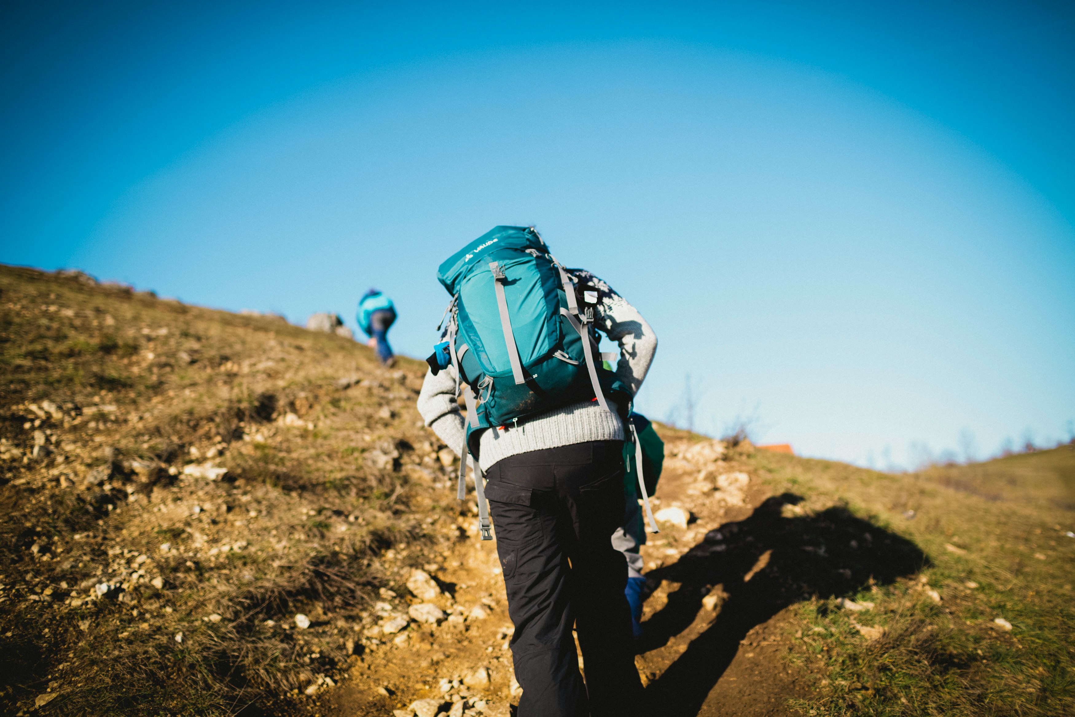 man in blue jacket and black pants with blue backpack walking on brown field during daytime