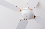 A close-up view of a white ceiling fan with four blades. The fan is mounted on the ceiling with a minimalistic design, featuring metallic screws at the base of each blade. The white color of the fan blends into the ceiling, providing a clean and simple aesthetic.